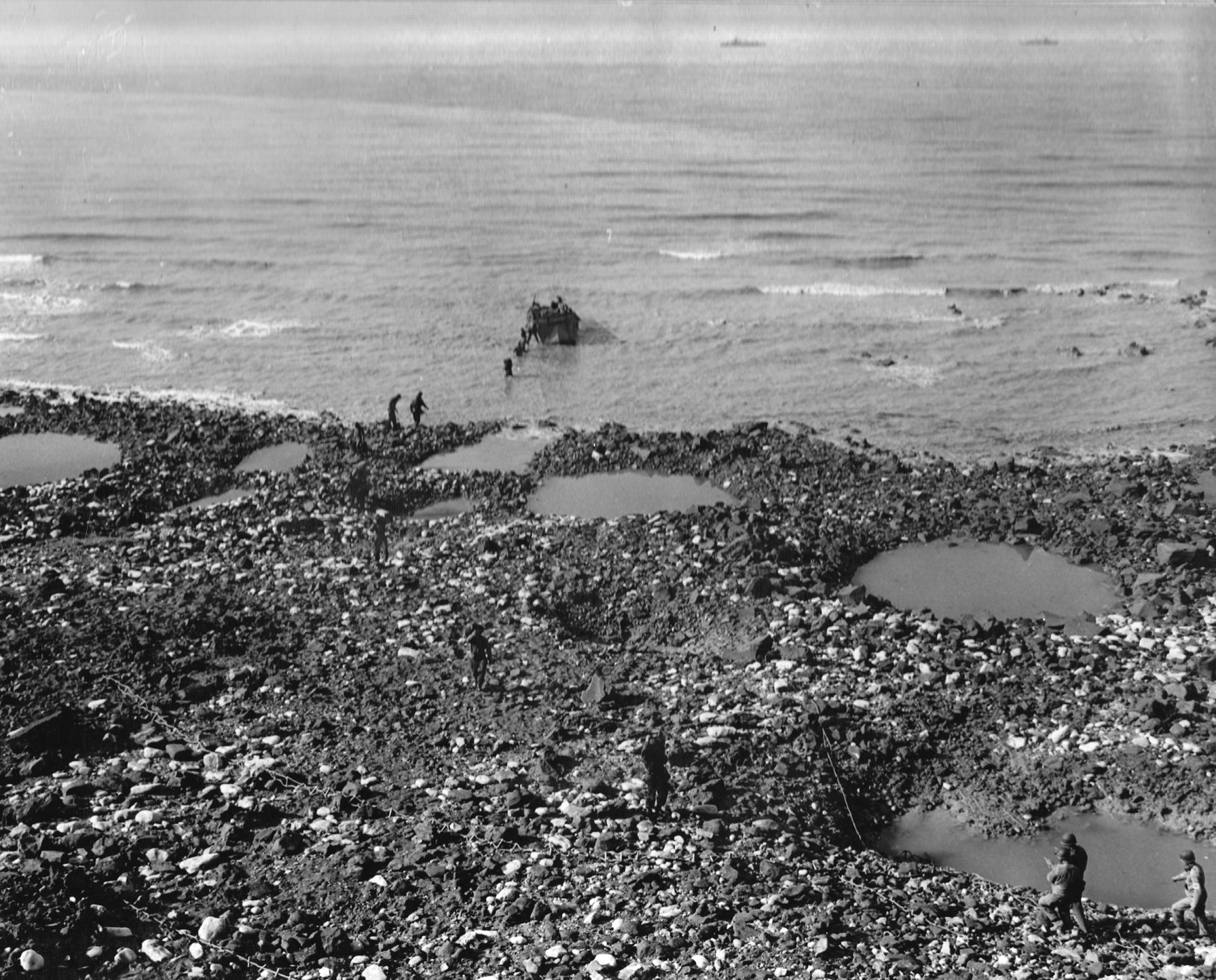 After Pointe du Hoc has been secured, a boat brings supplies ashore to beleaguered Rangers who have defended the position for hours. A pair of Allied naval vessels lies in the distance, and scaling ropes lay on the beach.
