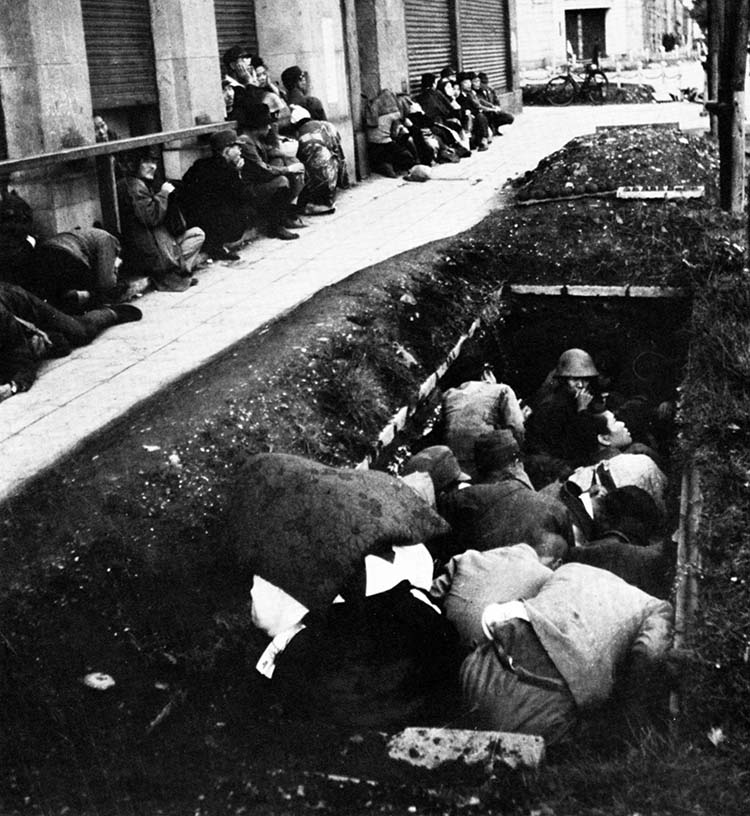 Fearing another large bombing raid by American aircraft, Japanese civilians in the Ginza District of Tokyo take cover in doorways and a recently dug trench as a U.S. reconnaissance plane flies overhead.