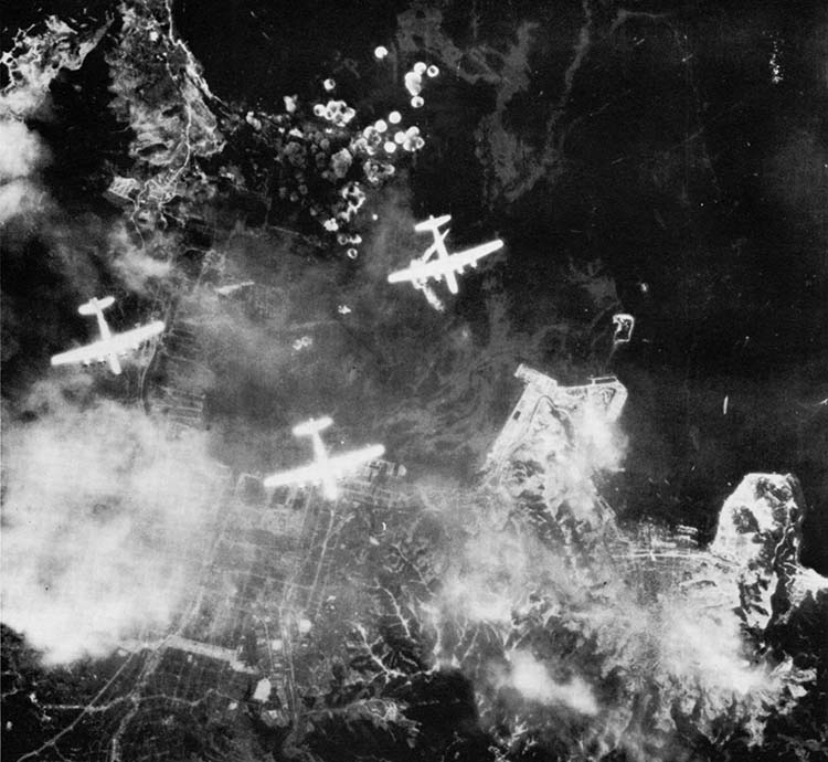 As smoke billows from the stricken Japanese naval base at Kure, American bombers fly high above and release another payload of destruction. Japanese cities and military installations in the Home Islands were laid waste from the air.