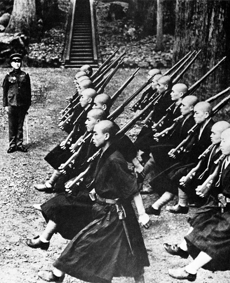 With rifles over their shoulders and bayonets lashed to their belts, Buddhist monks march in step under the watchful eye of a Japanese soldier. This drill took place on the grounds of their Buddhist temple.