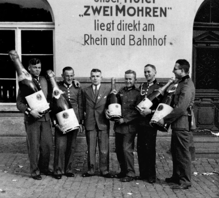 CELEBRATION: Dressed in their Waffenrock or “walking out” dress uniforms, a group of soldiers celebrates some occasion with extraordinarily large bottles of champagne. A clue to the photo’s strategic location is found on the wall behind them that reads, in part, “The Two Moors is situated directly on the Rhine and the railway station.” 