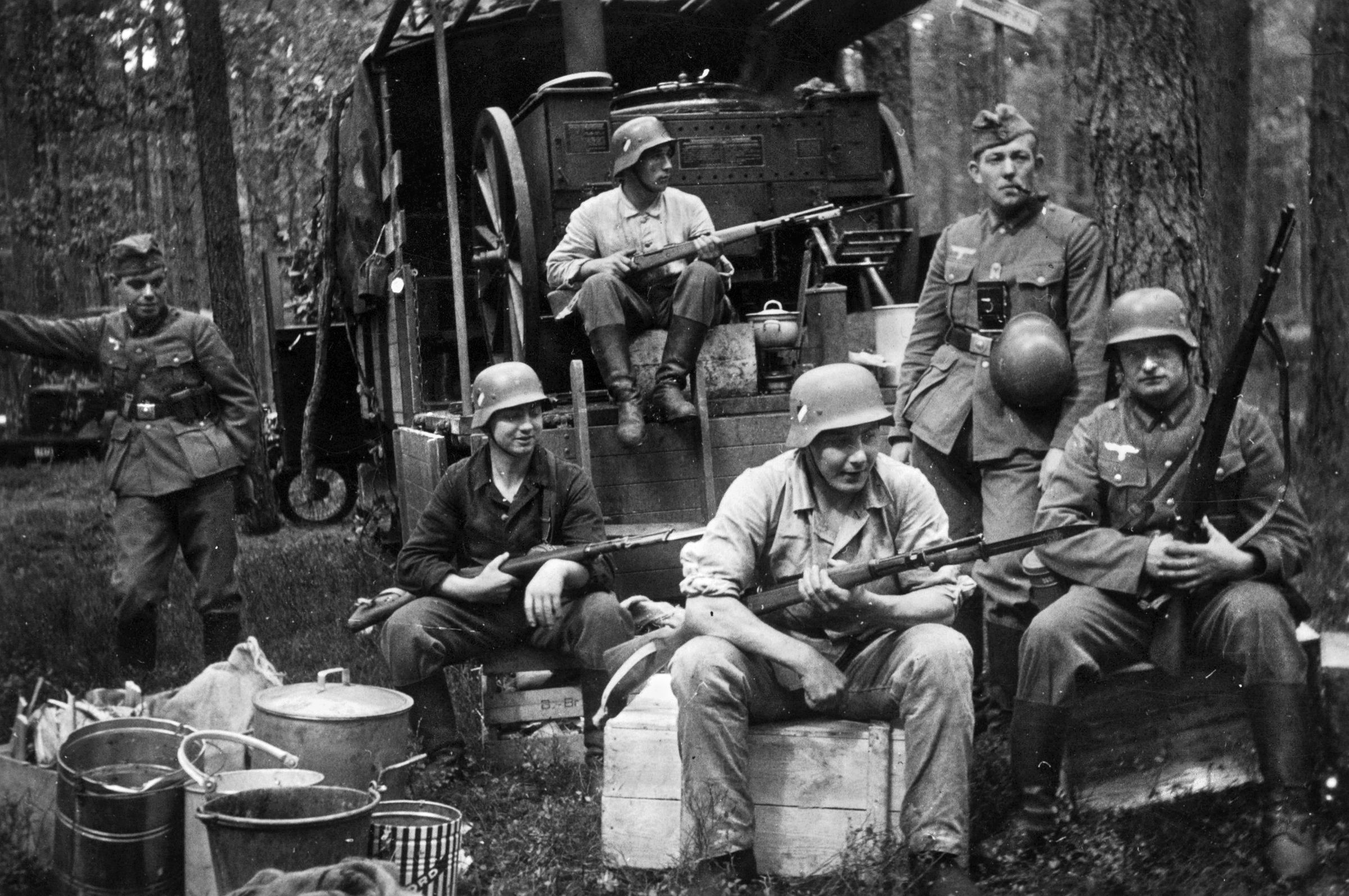 GUARDING THE KITCHEN: Bayonets at the ready, a contingent of soldiers exaggeratedly guard the all-important Feldküchen (field kitchen), seen transported within a larger wagon. Pulled either  by a motorized vehicle or by horse, mobile field kitchens or Feldküchennwagen incorporated a wood, coal, or charcoal-fed stove. The compact unit brought hot meals, usually stews or soup, to the troops in the field. One frequently seen item on the menu was Frontkameradensuppe, which consisted of a stew of beans, potatoes and ham (aka “the comrades”).   