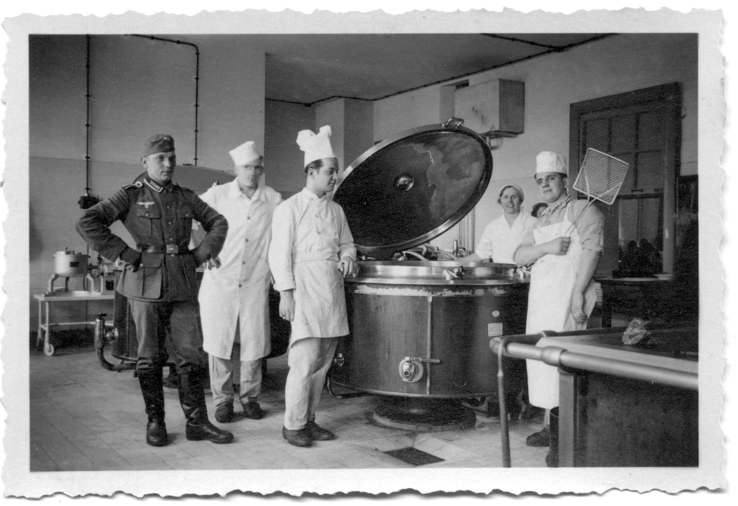 INDUSTRIAL DEEP-FRYER: Seen within a spic-and-span, state-of-the-art military base kitchen, the staff poses proudly by one of their massive cooking vats. A spigot is visible from which the grease is released into a floor trap for recycling. Grease was a component used in the production of explosives. 
