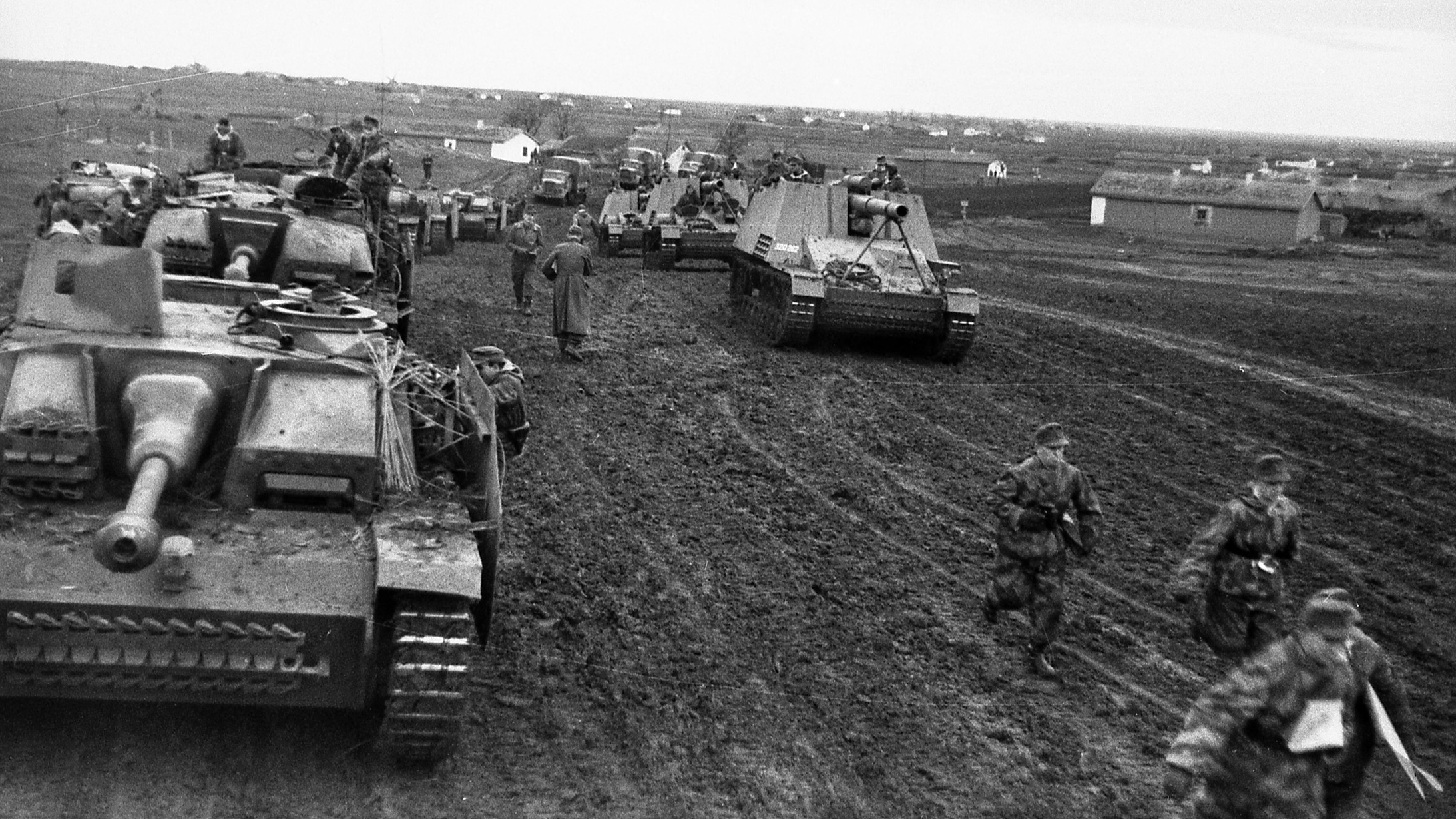 Traversing a muddy road on the outskirts of a Ukrainian village, two columns of German assault guns move toward the front lines. The vehicle to the left is the Sturmgeschütz III mounting a 75mm gun, and the vehicle on the right is the Hummel (Bumblebee) self-propelled 150mm howitzer.
