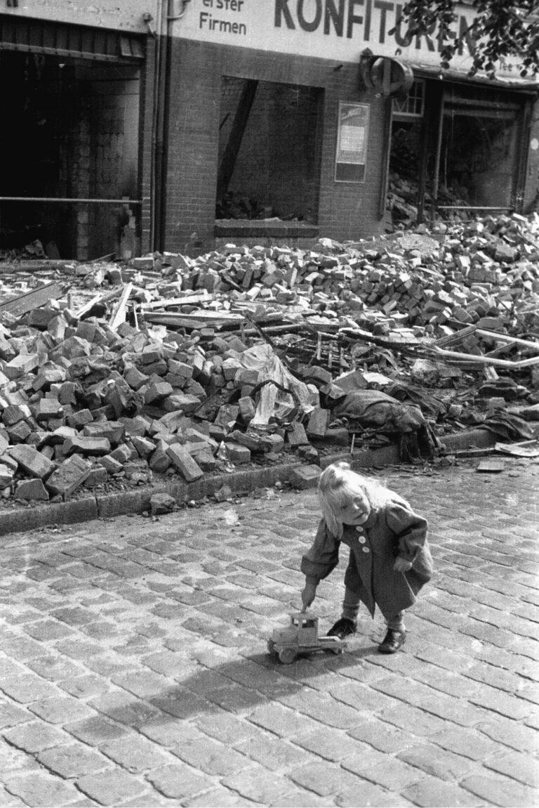 A lucky German girl with a warm coat and a rare toy truck plays in front of a pile of rubble in her bombed-out city.