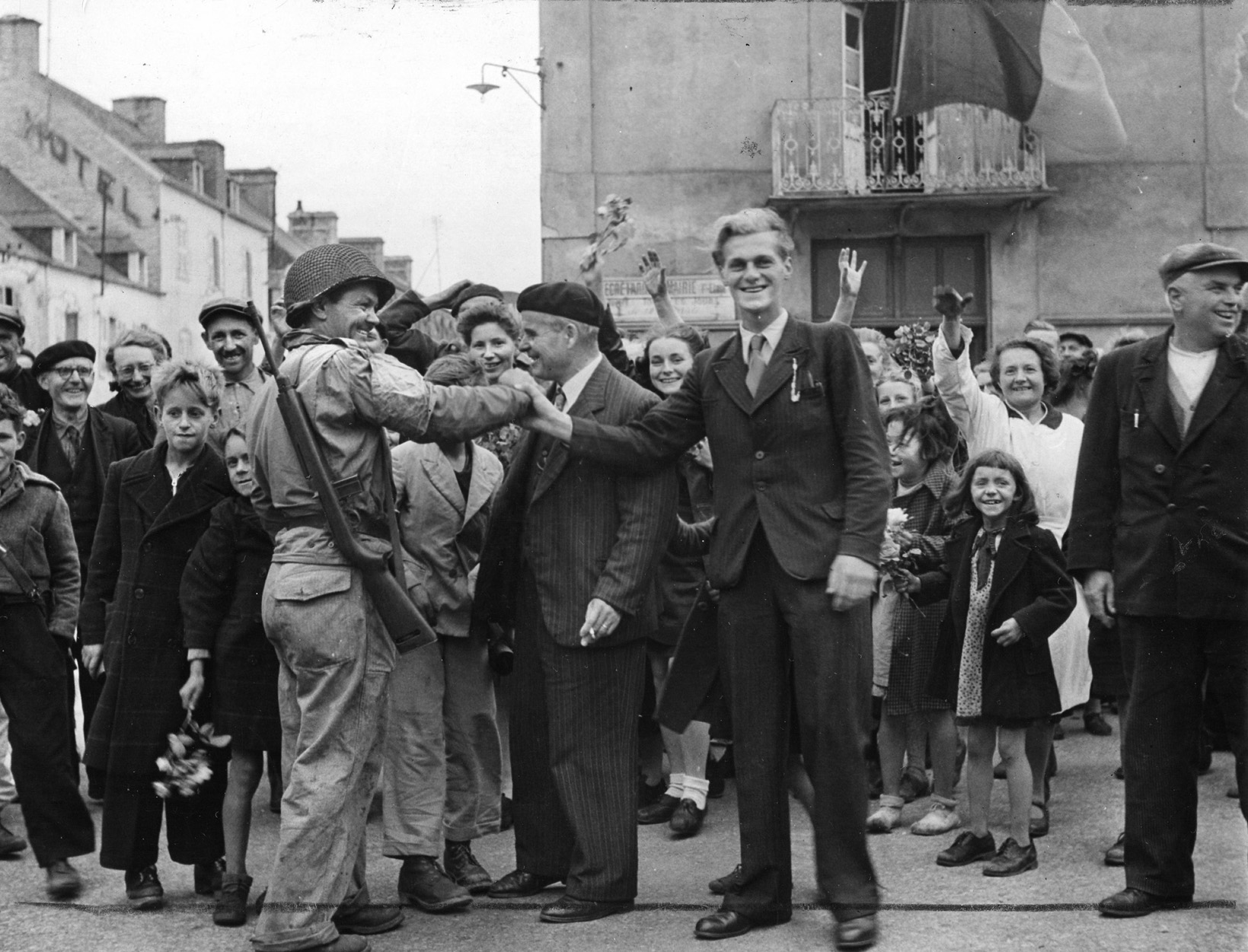 The mayor of Barneville, wearing a beret, greets an American sergeant while jubilant residents wave flowers.