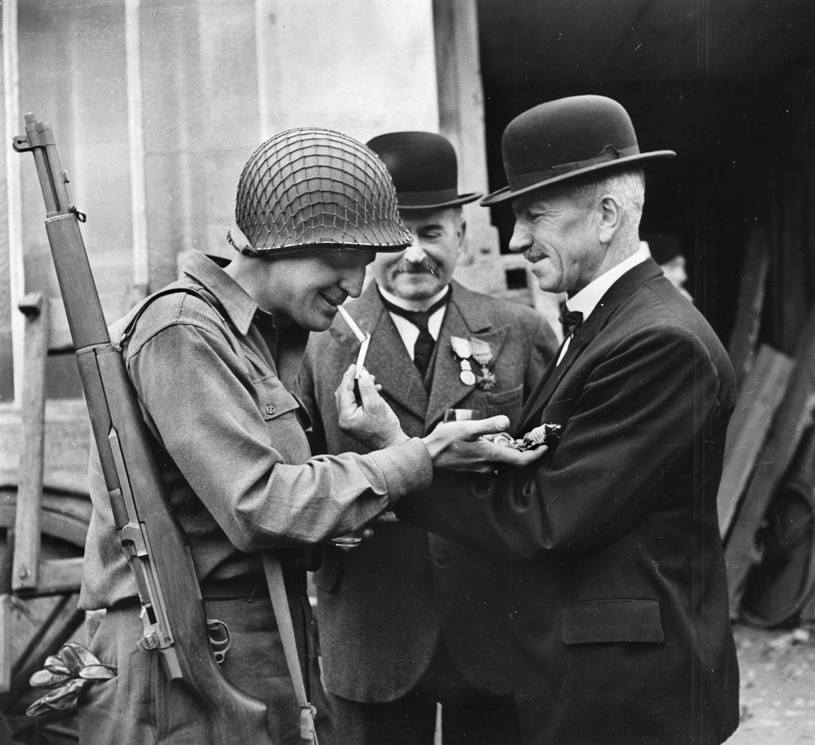 A GI examines a Frenchman’s World War I medals while the Frenchman offers a chain to the American’s cigarette.