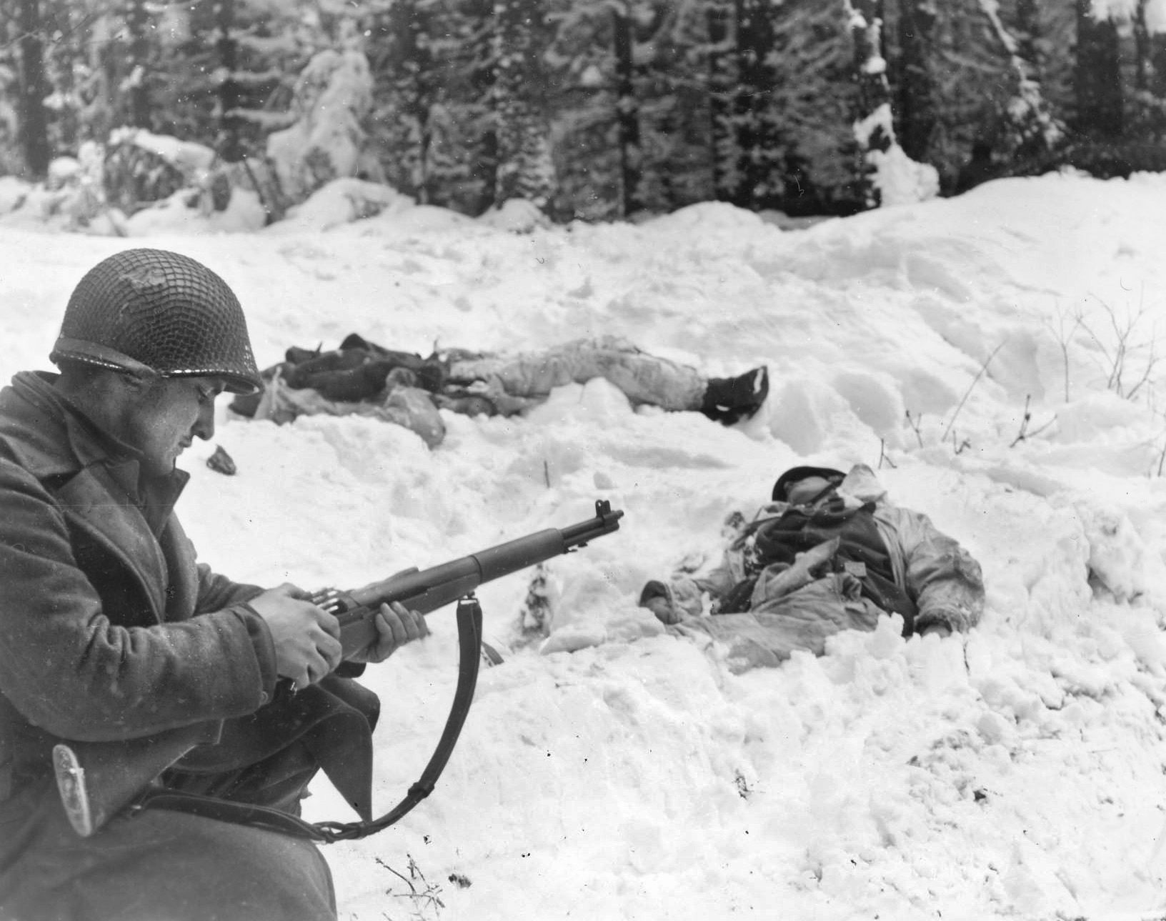 A GI calmly loads his M-1 Garand near two dead Germans whose white camouflage clothing did them no good. 