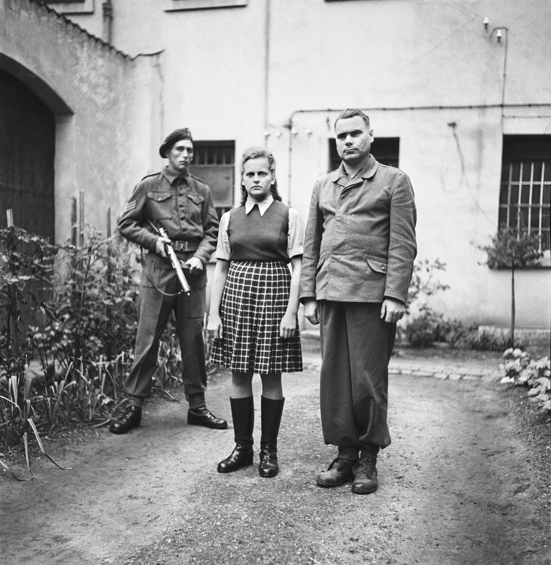 In jackboots and civilian clothes, Irma Grese (along with Bergen-Belsen commandant Josef Kramer, right) stands in the courtyard of the prison at Celle during their war-crimes trial, June 1945.