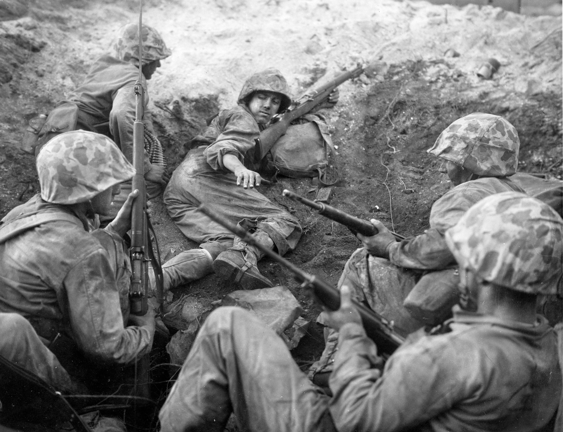 A U.S. Marine squad leader motions to his men to stay low within the protection  of a shell hole on the island of Namur. The Marines assaulted Namur and the island of Roi on February 1, 1944, with a primary focus on seizing the airfield that took up nearly the entire landmass of Roi. Preinvasion bombardment had given the Marines plenty of craters  in which to take cover.