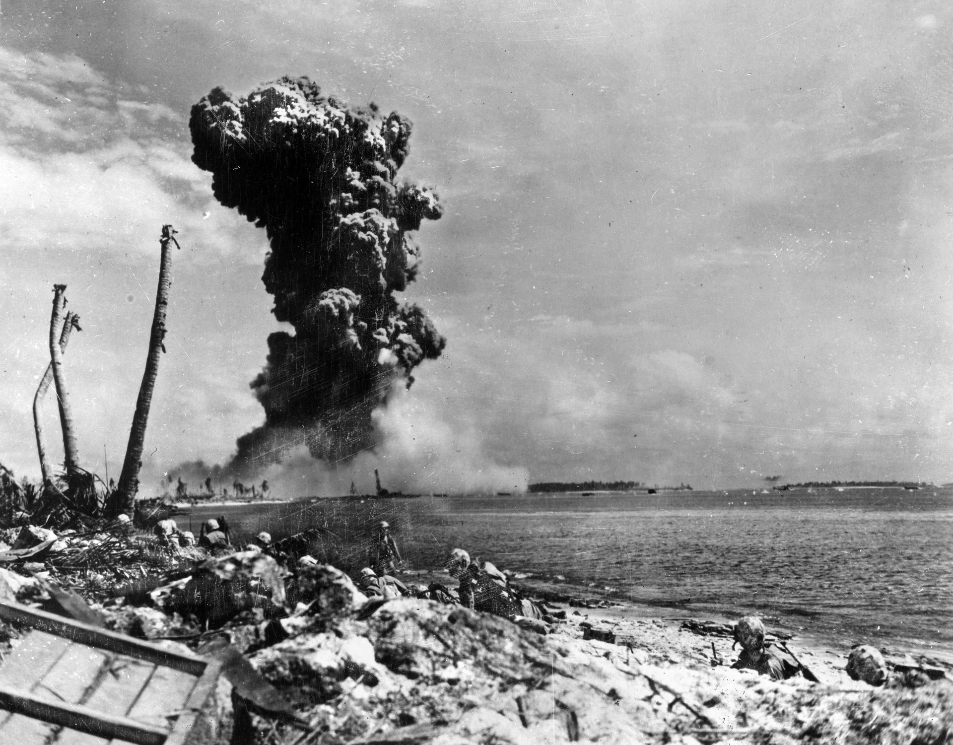 Having taken cover on the beach at Roi, American Marines look across the lagoon toward Namur as a huge explosion destroys a Japanese bunker. Marines on the adjacent island had placed a satchel charge to destroy the bunker, and its demolition ignited an ammunition storage area with spectacular results.