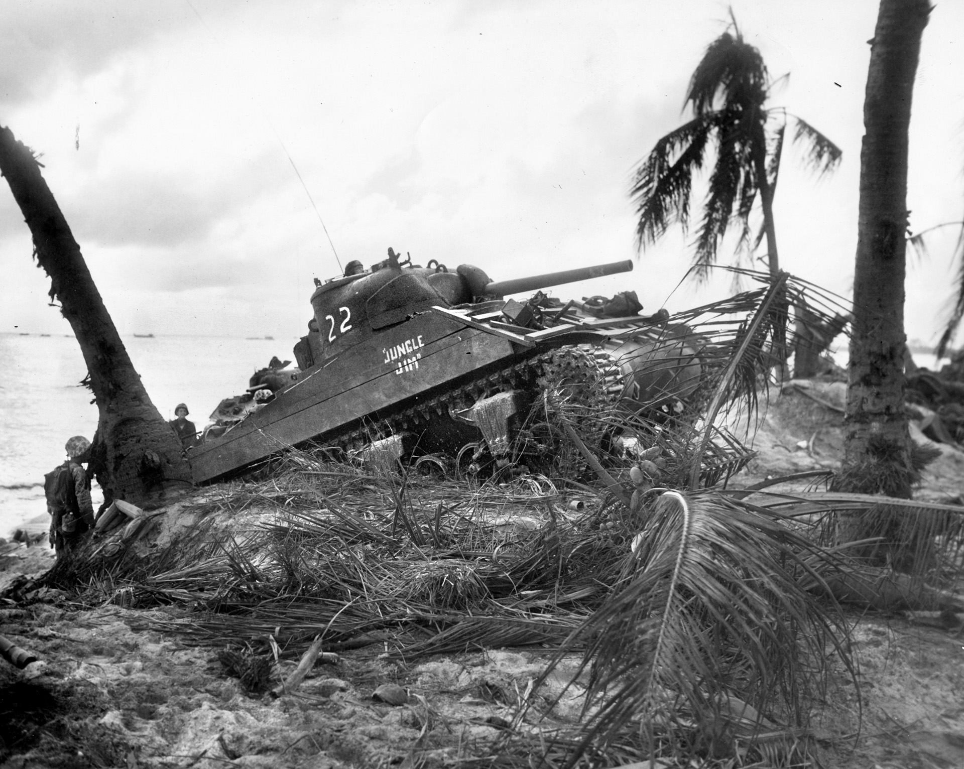 Having taken cover on the beach at Roi, American Marines With a couple of Marines watching intently, an M4 Sherman medium tank moves across the sandy berm adjacent to the beach at Namur.