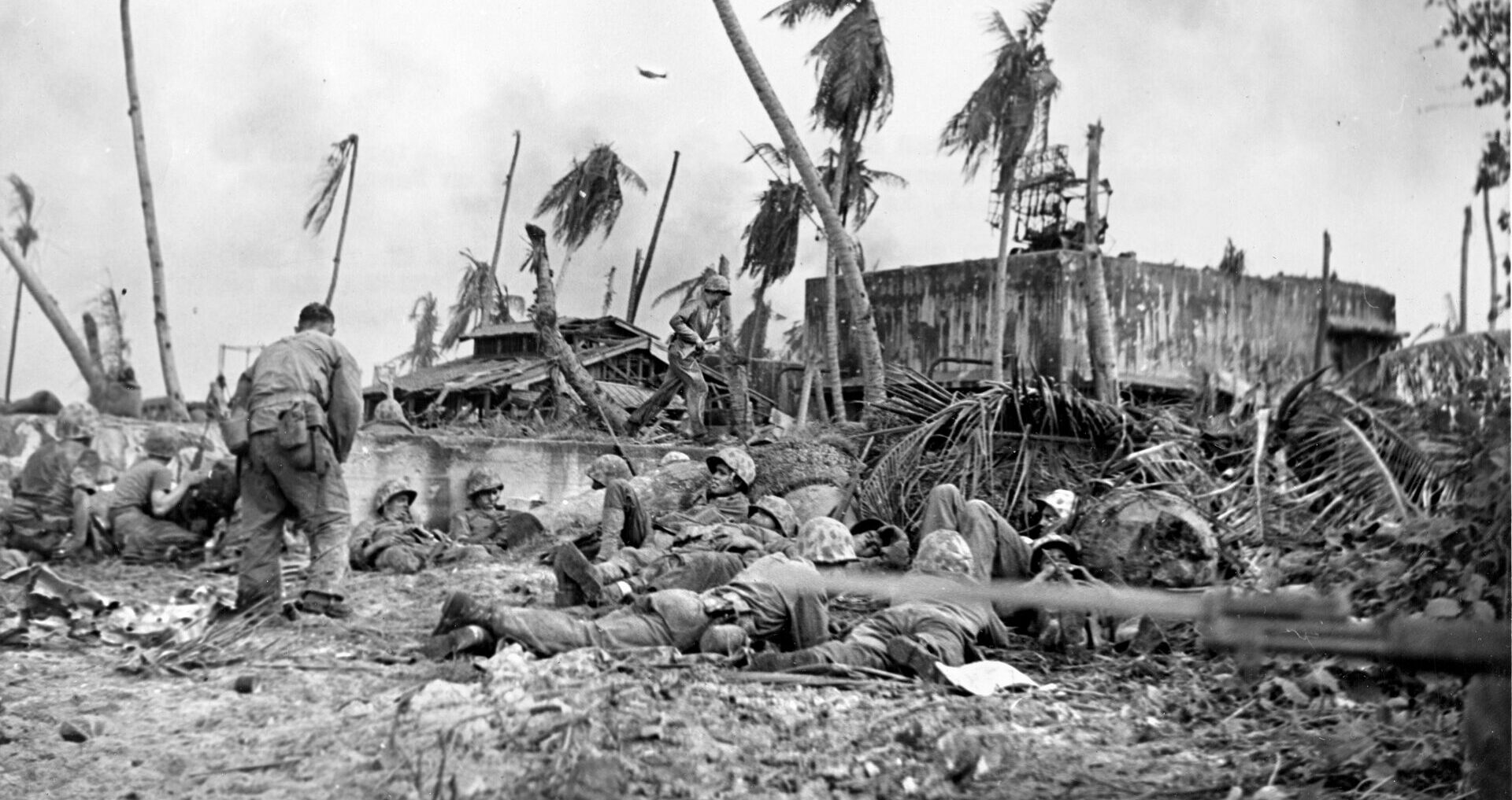 The defending Japanese had heavily fortified an administration building on Namur, and these American Marines are taking cover during operations to silence enemy rifles and machine guns firing at them from its windows and apertures. An American aircraft flying in support of the ground troops is visible in the distance.