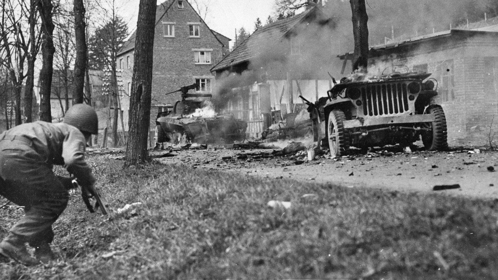 An American soldier cautiously approaches two burning vehicles that had been destroyed by a German ambush. As a scout, Private Sevel never wore equipment or heavy clothing in order to stay mobile on the battlefield.