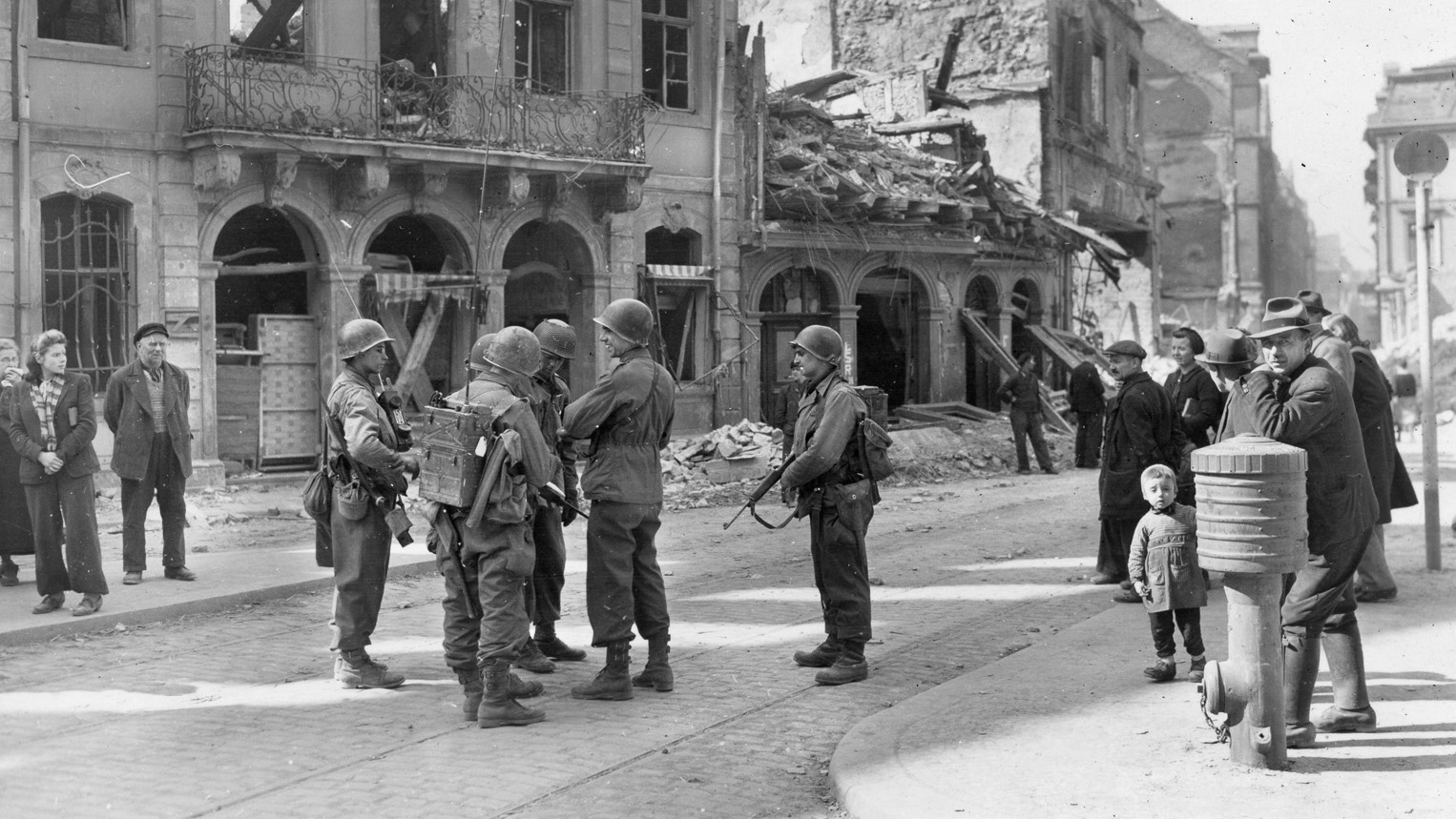 As German civilians look on, soldiers of the 90th Infantry Division’s 359th Infantry Regiment discuss their battle plans in the German city of Mainz.