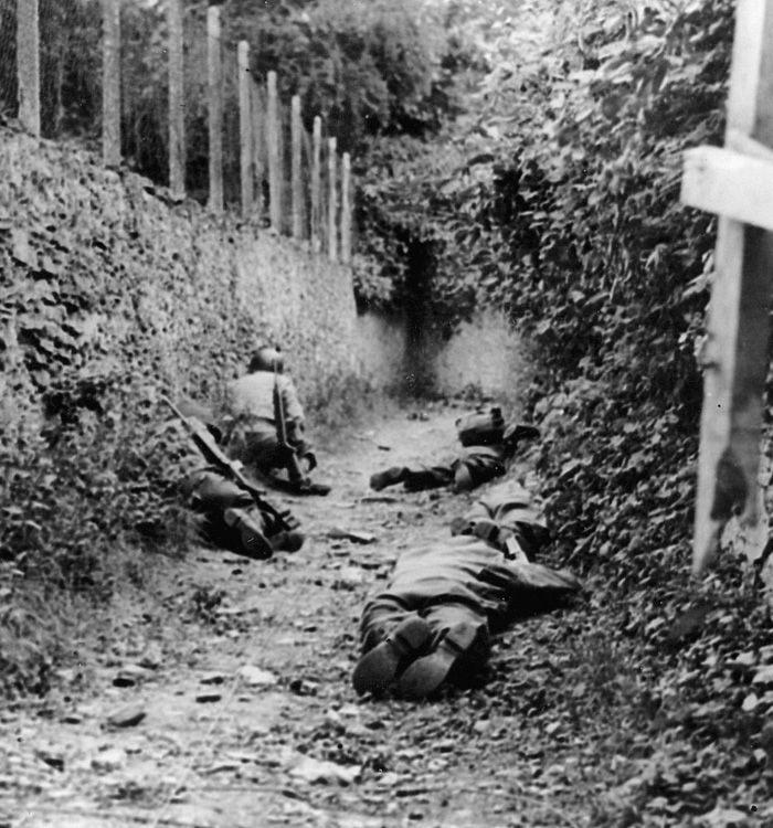 Crouching low against a wall and the earthen mound of a French hedgerow, U.S. soldiers attempt to avoid German artillery fire overhead.