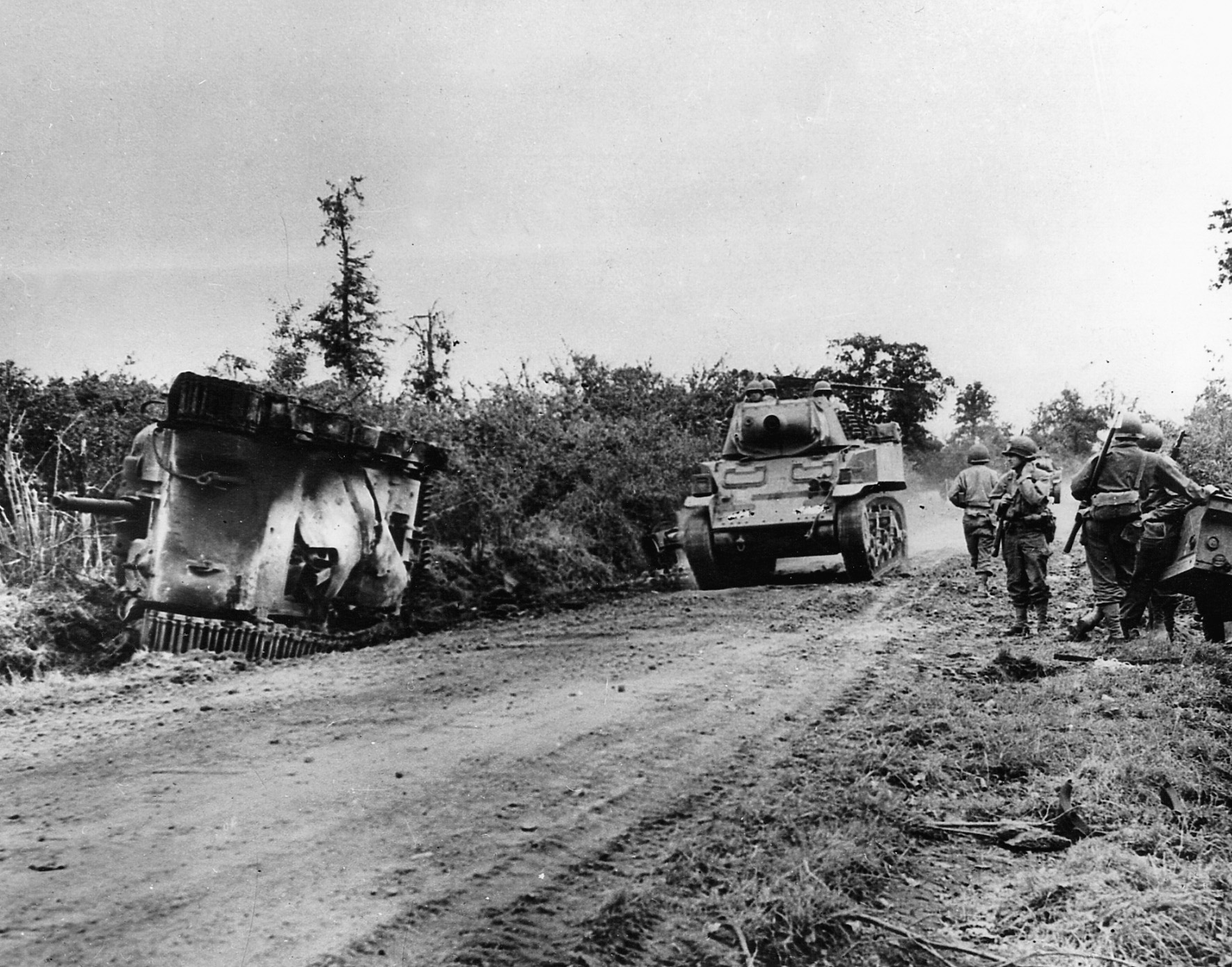 Passing the charred remains of one of its own, a U.S. Stuart light tank makes its way down a dusty road towards St. Lô. 
