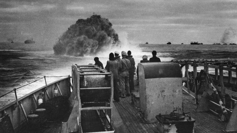 Crewmen aboard the U.S. Coast Guard Cutter Spencer watch the waters of the Atlantic Ocean brew up with the detonation of a depth charge. This photograph was taken while the Spencer was defending a trans-Atlantic convoy, visible in the background, against a German U-boat attack.
