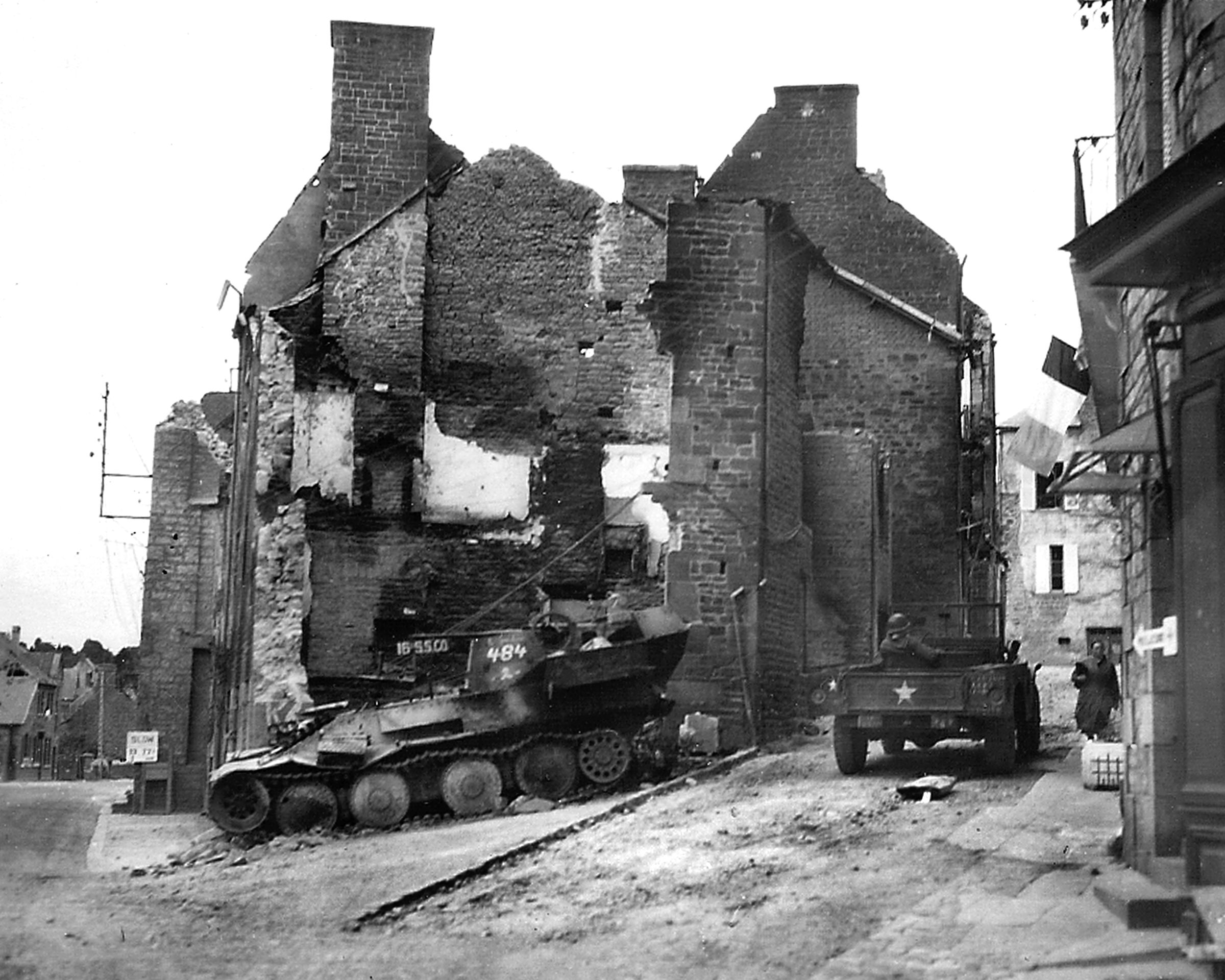 An American three-quarter-ton Dodge weapons carrier WC-51 passes a destroyed German armored vehicle, a victim of the fierce fighting in Carentan several weeks earlier.