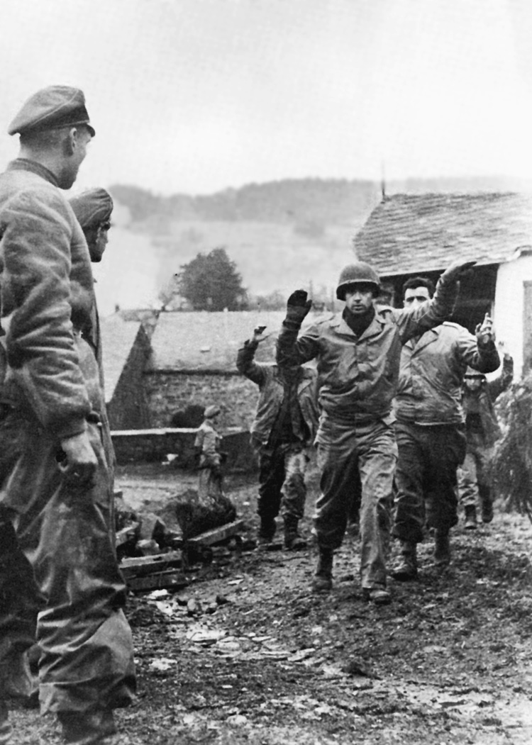 During the early phase of the Battle of the Bulge, advancing German SS troops overwhelmed forward American positions in the Ardennes Forest. In this photo, an American prisoner gestures toward a group of SS officers to ask if he and his fellow captives are marching in the right direction.