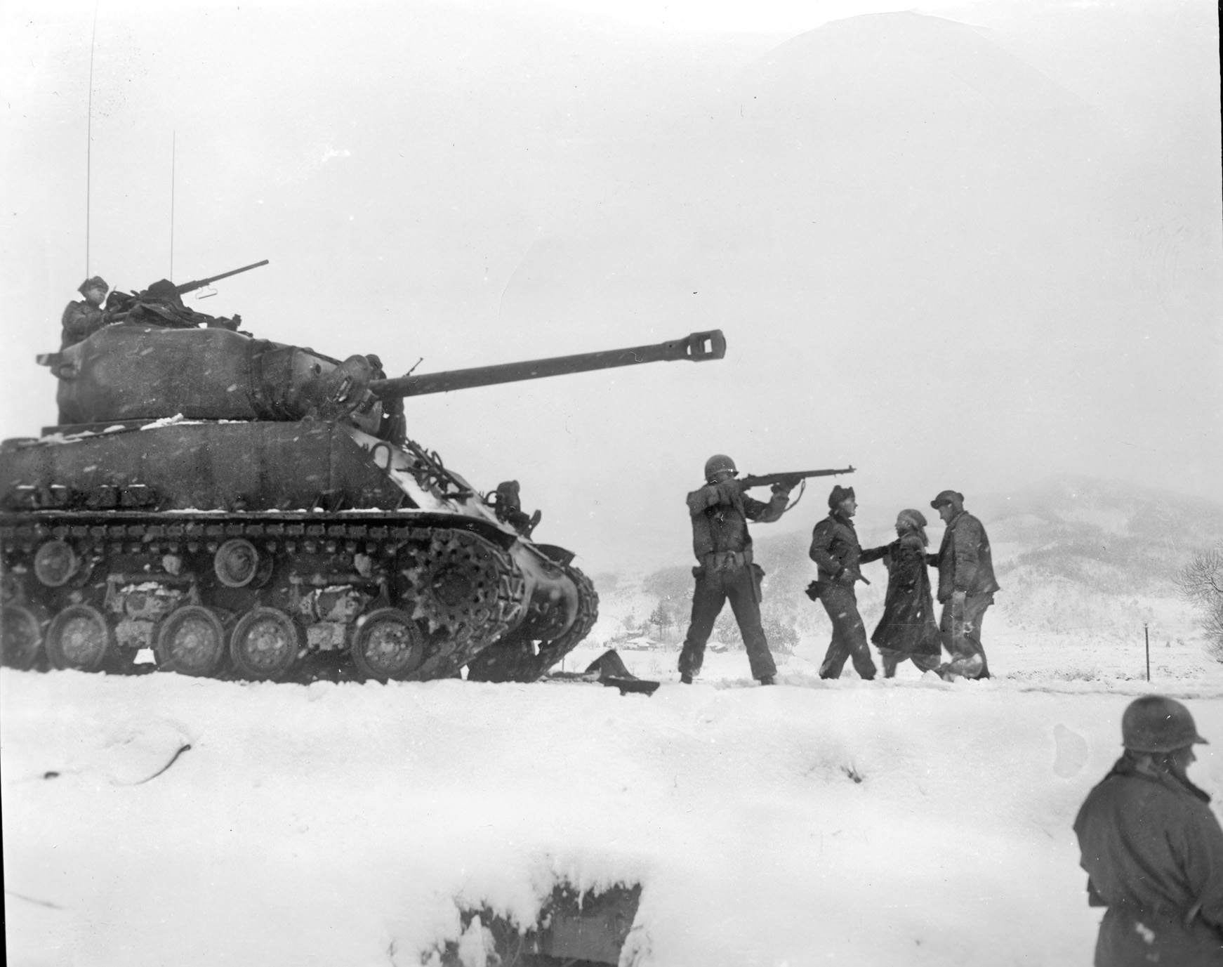 A tank of the Heavy Tank Company, 7th Infantry Division, engages the enemy at the Chosin Reservoir as infantrymen escort a prisoner to the rear.