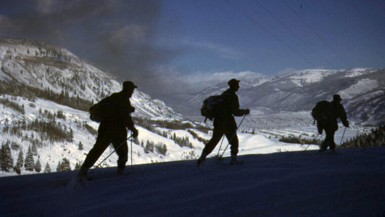Three 10th Mountain Division soldiers ski above Camp Hale and the Pando Valley. A cloud of dark coal smoke from stoves and passing freight trains darkens the otherwise pristine mountain air.
