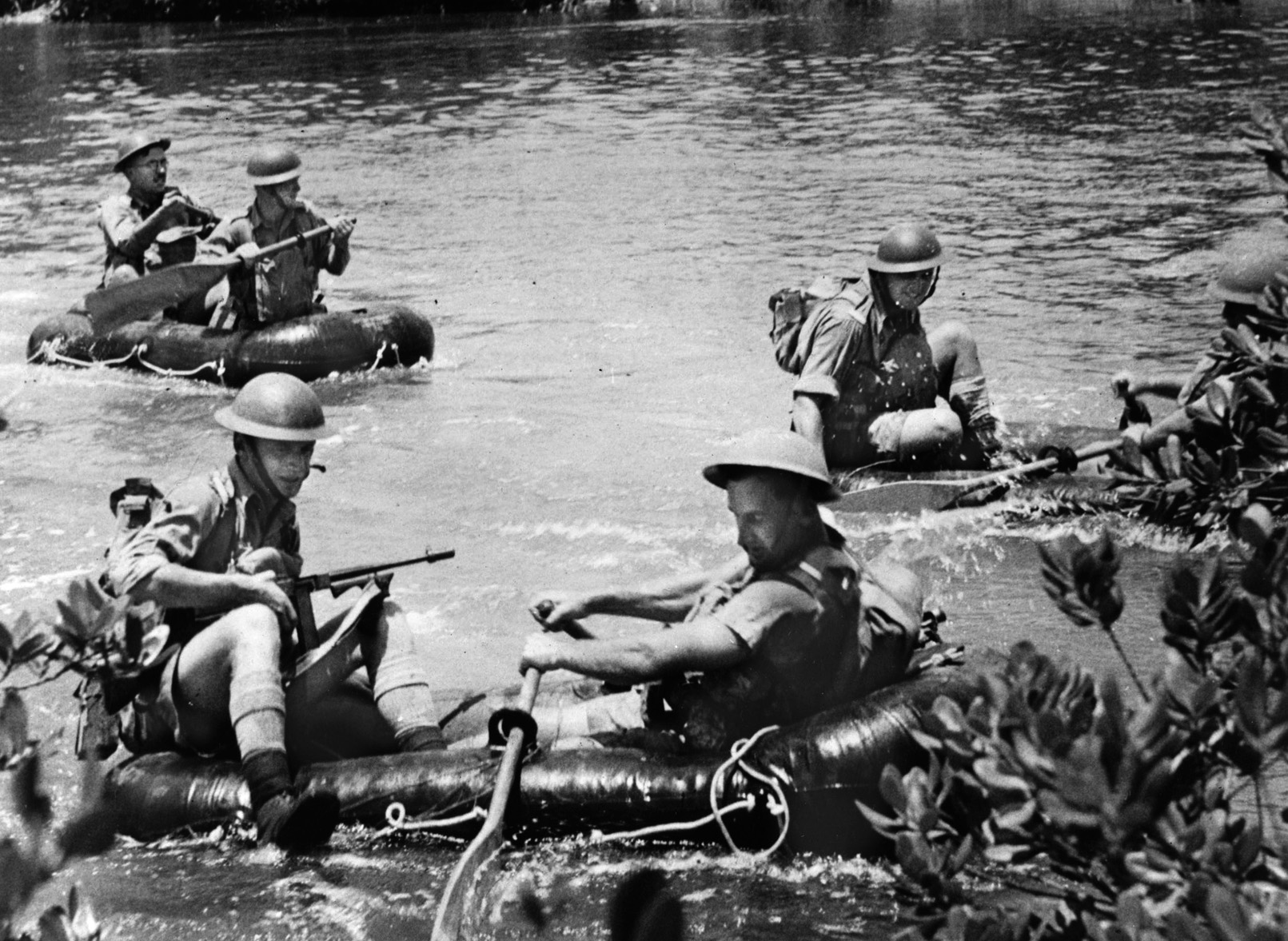 Six Australian soldiers with tommy guns cross a jungle river in rubber rafts during training. The Aussies worried that if Malaya fell, Australia probably would be invaded next.