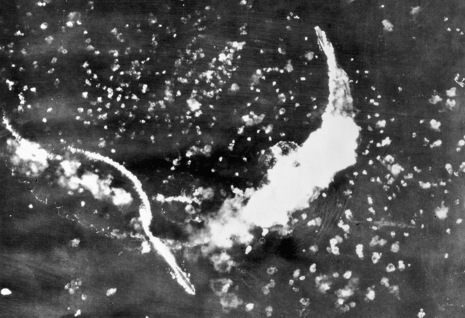 Surrounded by bomb bursts, a carrier throws up a smoke screen during an attack by enemy aircraft. 