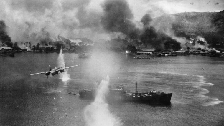 A pall of black smoke hangs over the shore installations at Rabaul as a B-25 medium bomber streaks above a Japanese merchant ship riding at anchor.