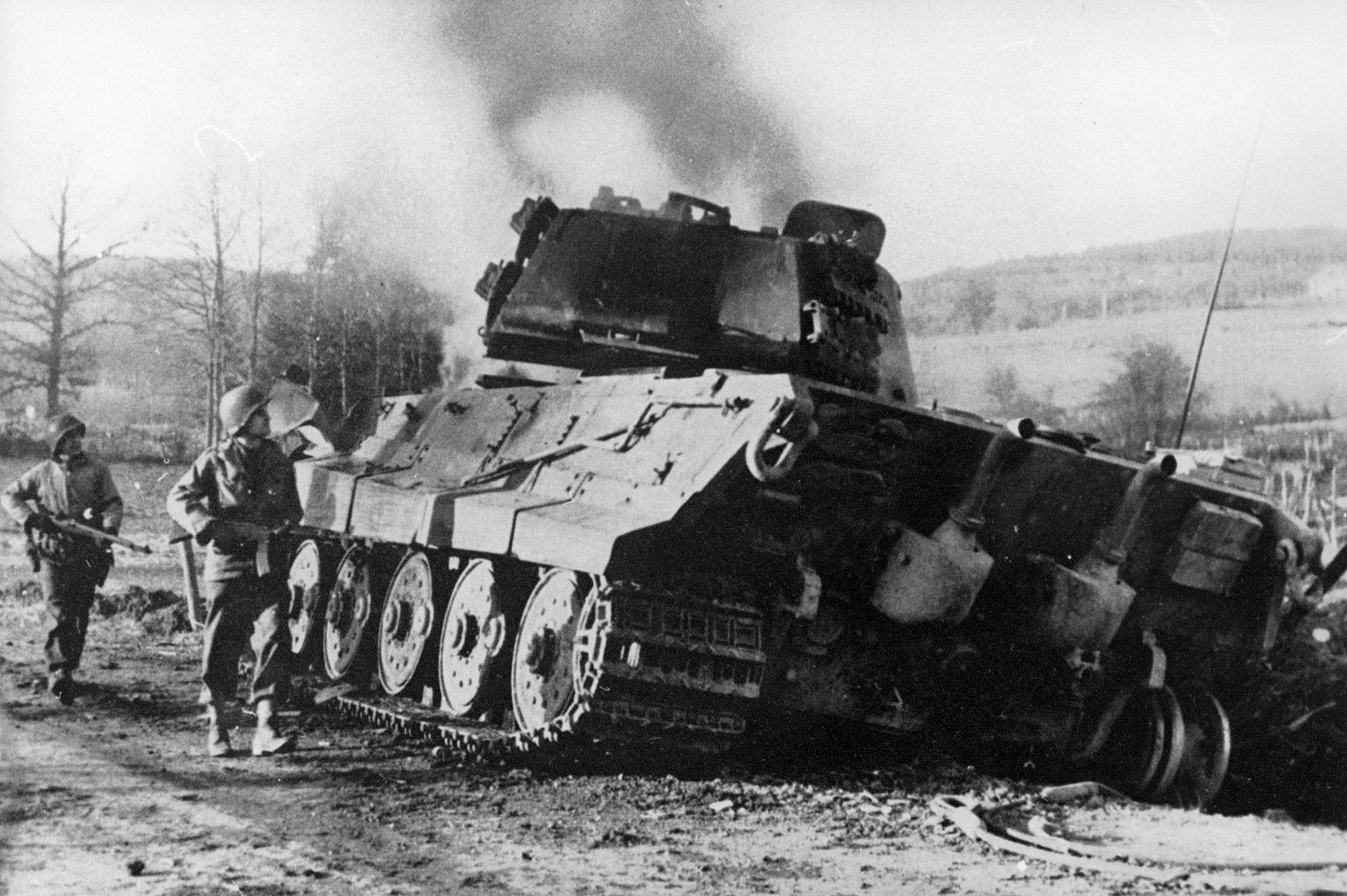 Two American soldiers advance down a muddy road near the Belgian village of La Gleize and glance at the smoking hulk of a German Panther medium tank that has recently been destroyed. 