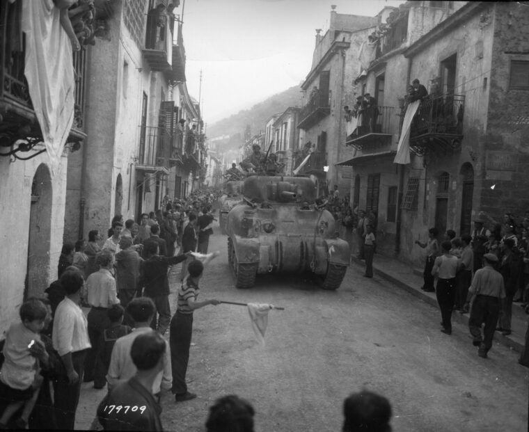 Tankers of the 2nd Armored Division roll down one of Palermo’s narrow streets while civilians cheer.