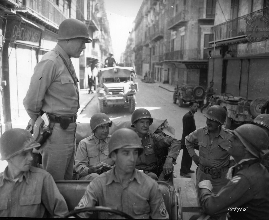 Lieutenant General George S. Patton, with one of his ivory-handled pistols on display, discusses the capture of Palermo with Maj. Gen. Lucian Truscott, who is wearing the blue-and-white striped patch of his 3rd Infantry Division. Maj. Gen. Geoffrey Keyes, who commanded the Provisional Corps, sits in the left seat of Patton’s command car.