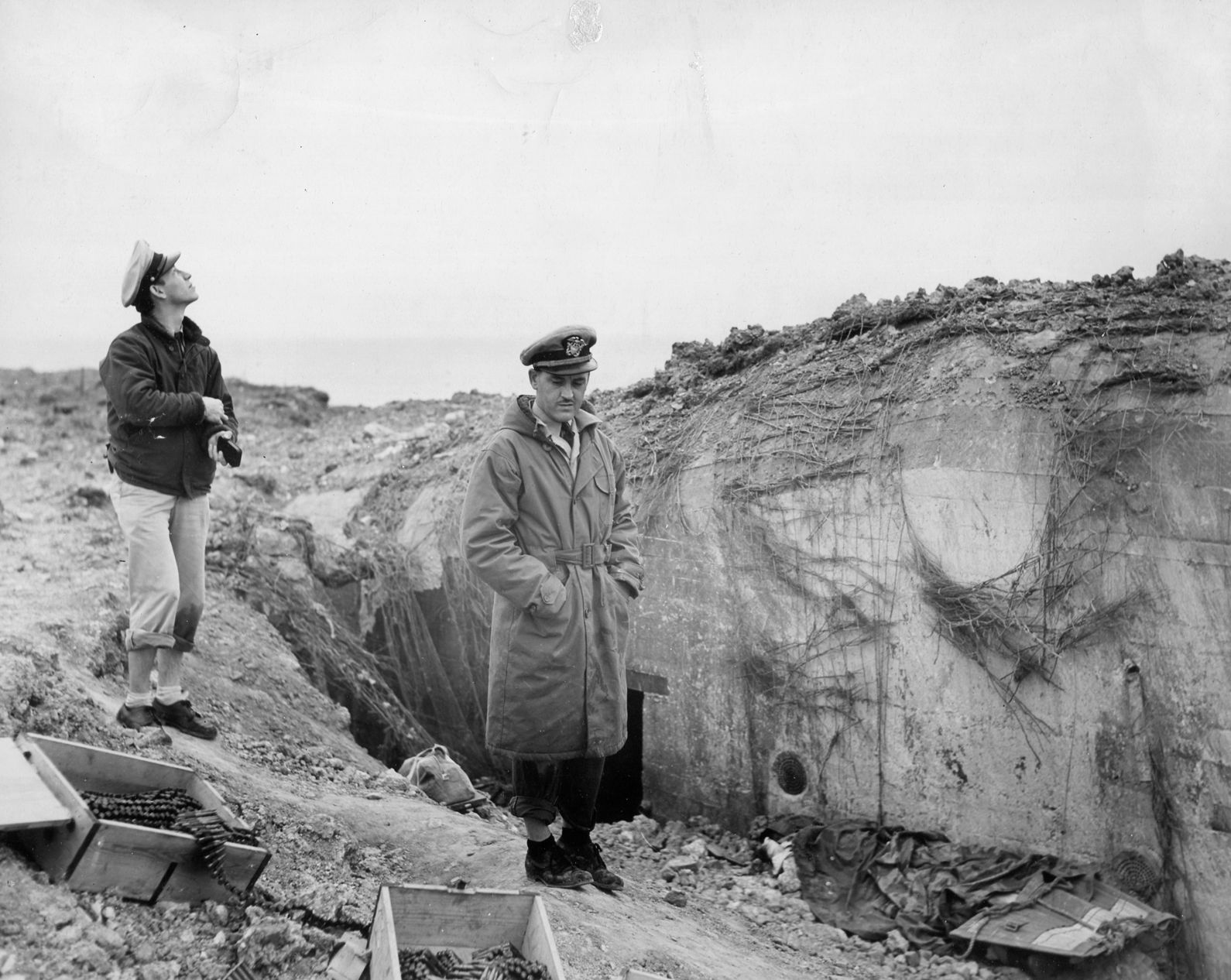Two Navy officers from USS Texas examine a German pillbox atop Pointe du Hoc. A box of machine-gun ammunition is at lower left, while a dead soldier, covered by blankets, lies at bottom right. 