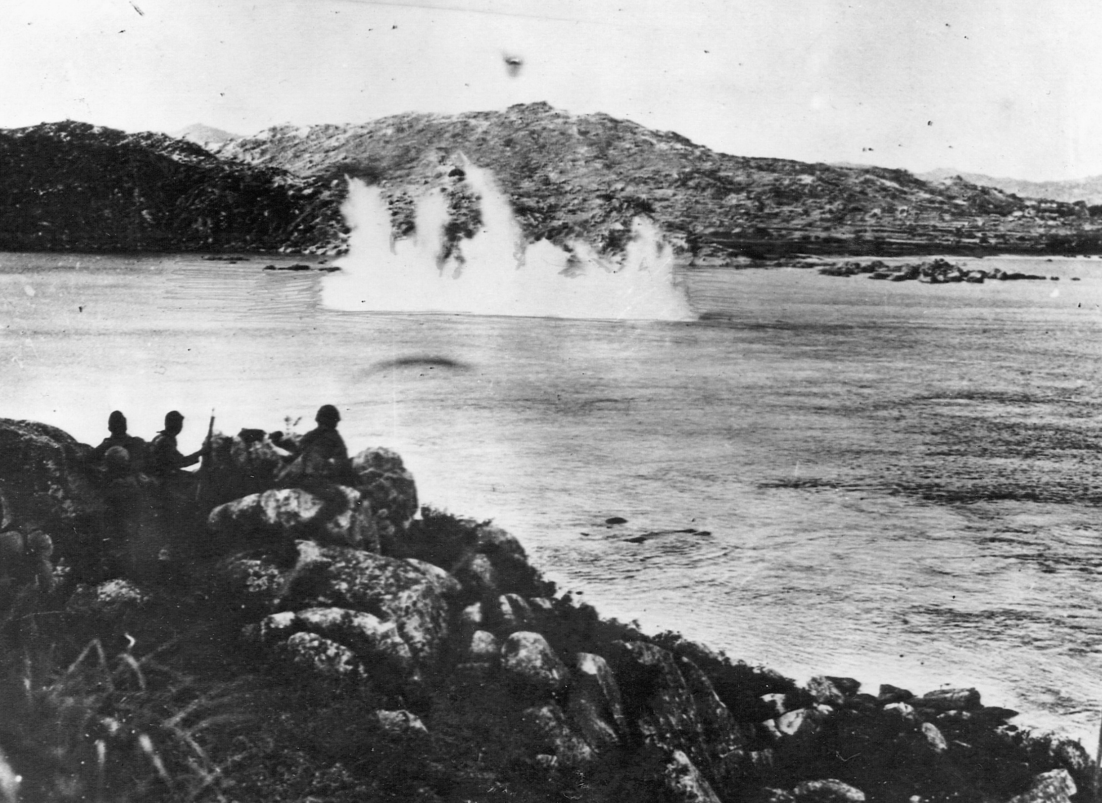 As a group of Japanese soldiers watches from ashore, a detonated mine sends plumes of water from the Yangtze River.