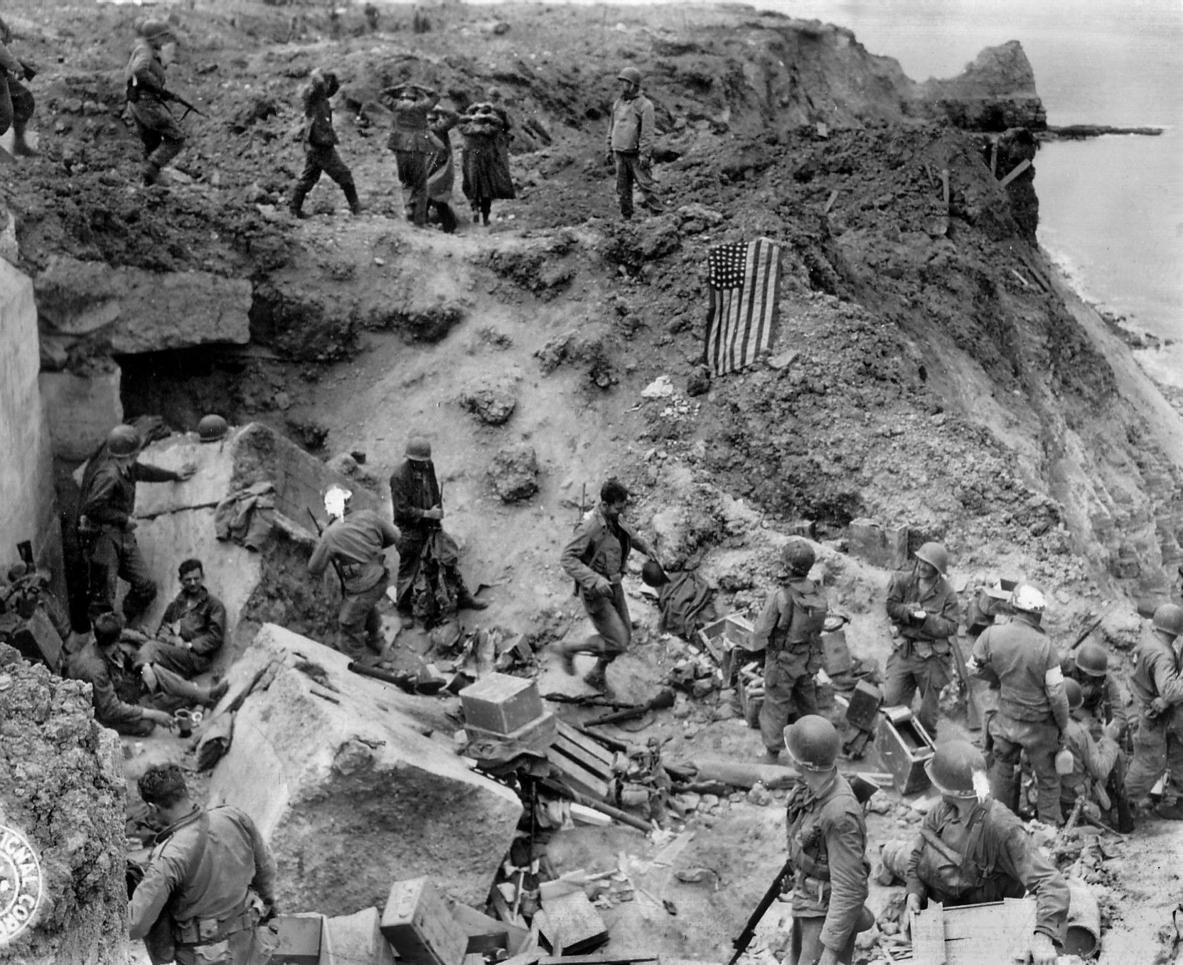 Rangers relax or get medical treatment at Lt. Col. Rudder’s rough-hewn command post while German prisoners march past Rudder, top right. Below him, the U.S. flag is laid out to let Allied ships know that the area is under American control.