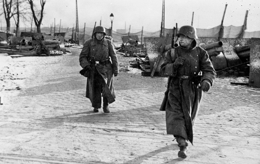 German sentries guard an area that is apparently in use as a depot for captured military equipment. They are stationed along the road near a bridge across the Warthe River near the city of Neustadt.