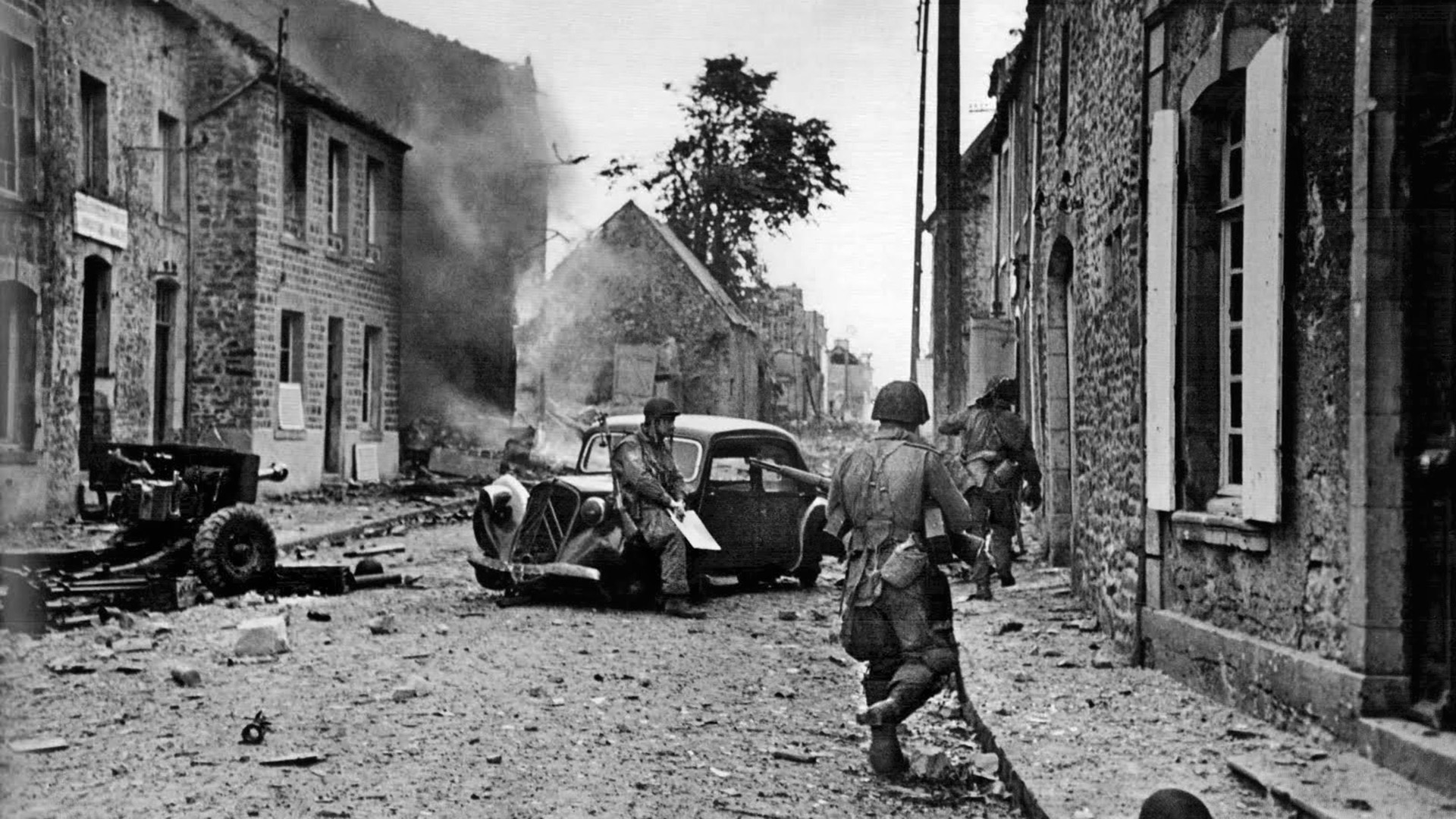Disabled automobiles lie abandoned in the street of a French village south of the major port city of Cherbourg. In this image soldiers of the U.S. 82nd Airborne Division move cautiously through the area.