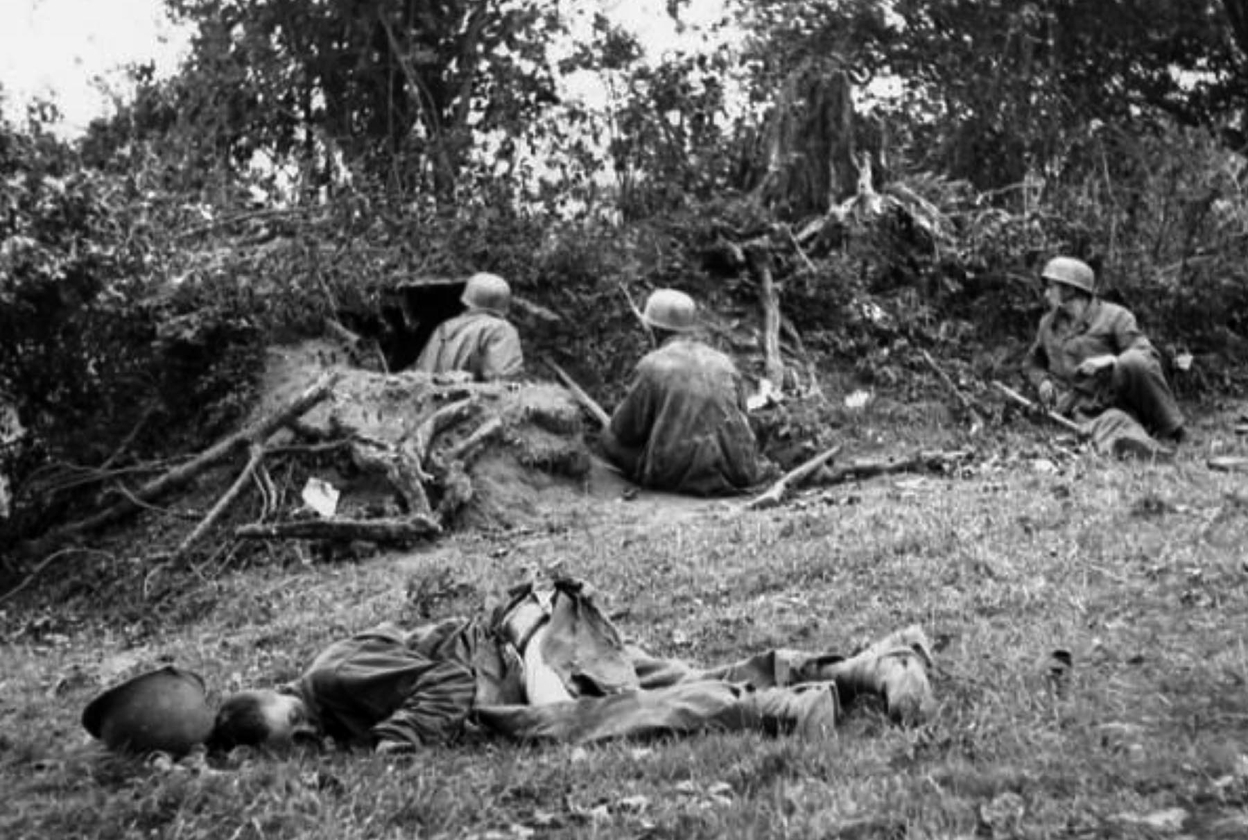 With the body of a dead American soldier lying nearby, three elite German paratroopers wait quietly in a well concealed defensive position along a Norman hedgerow. German machine guns and artillery were often pre-sighted to catch American and British troops as they came into view at the edge of a hedgerow.
