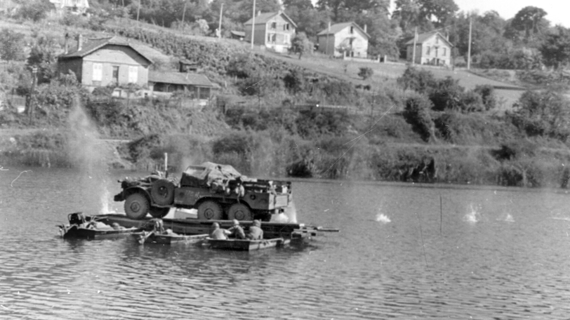 American combat engineers push a vehicle across the Seine aboard a float. Their heads are low as the crossing comes under fire from a German machine gun. Note the splashes of bullets hitting the water to the left and right. 