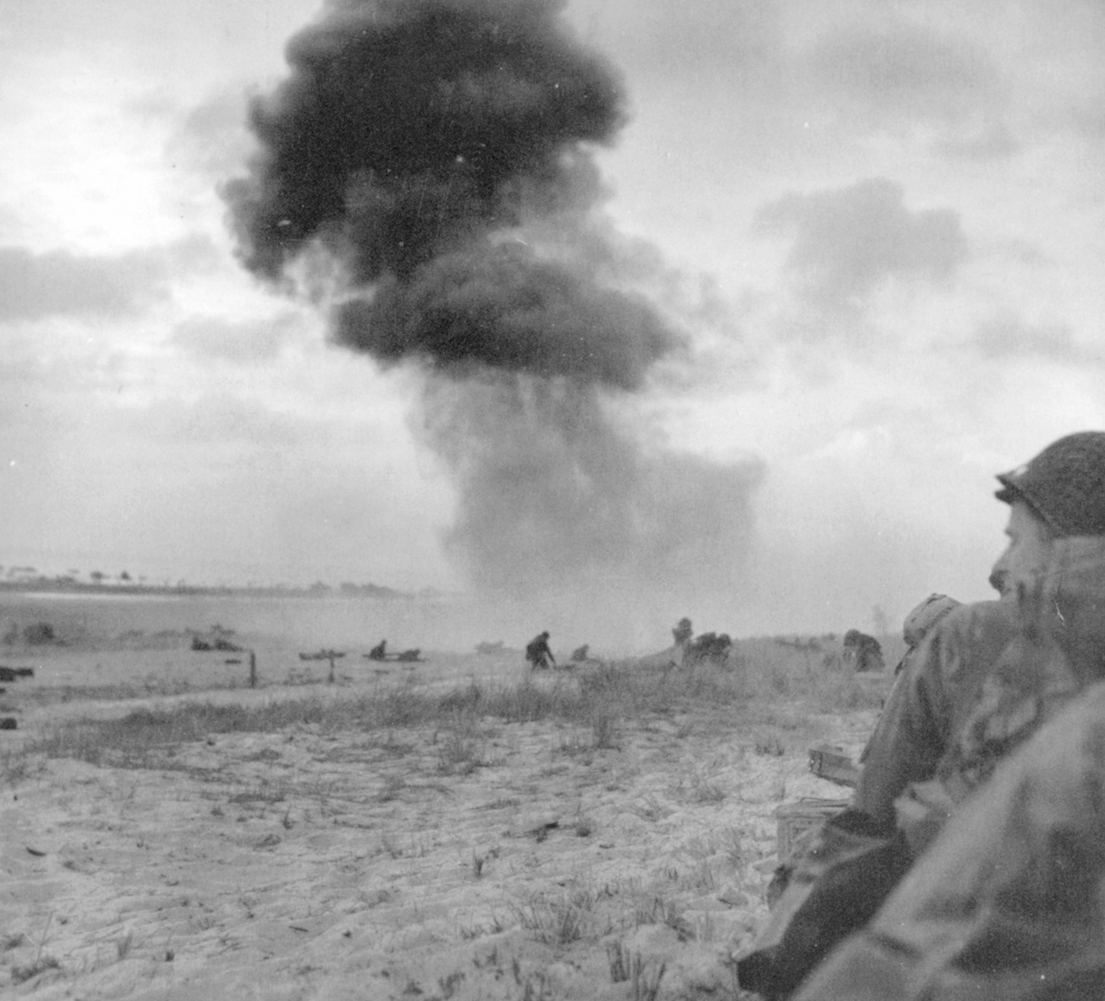  A soldier crouches for protection in the foreground while those in the distance scramble for cover as a German shell explodes on Omaha Beach.