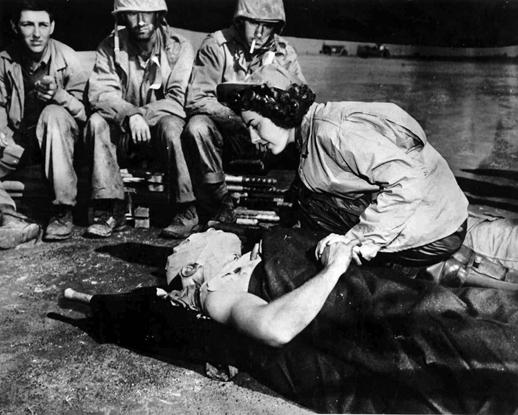 Worried buddies look on as Ensign Jane Kendeigh, a Navy flight nurse, comforts a badly wounded Marine before a flight out of Iwo Jima 12 days before the island was declared secure. Ensign Kendeigh was the first flight nurse on both Iwo Jima and, later, on Okinawa.
