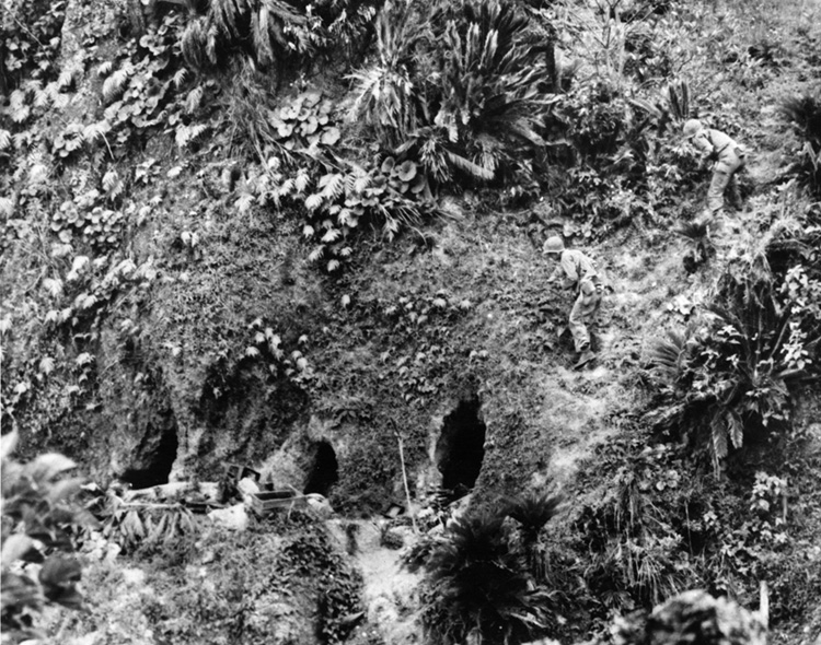 U.S. Army soldiers cautiously approach the mouths of two hillside caves with explosives in hand in order to kill enemy soldiers who refused to surrender.