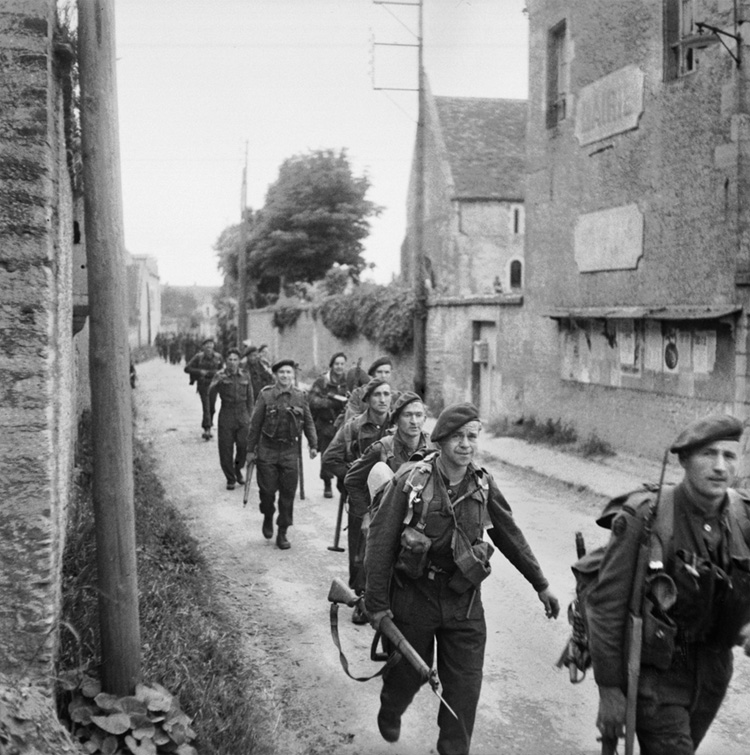 Photographed not long after landing on D-Day, these Royal Marine Commandos march through the town of Colleville-sur-Orne en route to the bridge and the relief of the lightly armed airborne soldiers.
