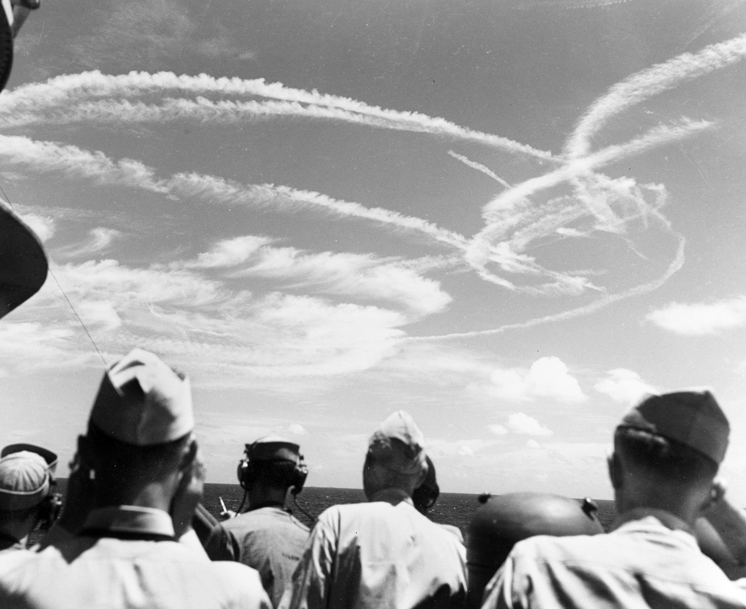 American naval personnel watch the action as white contrails criss-cross the skies while American fighters engage attacking Japanese aircraft. 