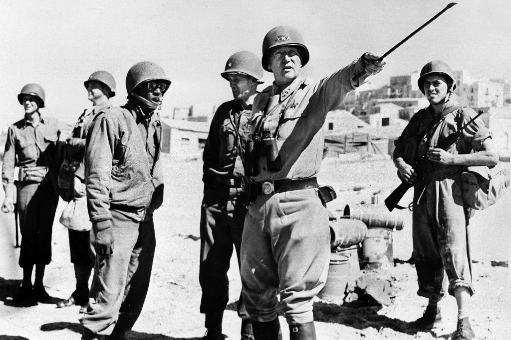 General Patton points to a distant objective while standing on the beach at Gela, Sicily, in 1943.   His aide, Sergeant William Meeks, stands to the general’s left.