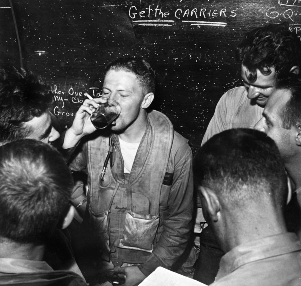 In the ready room of the aircraft carrier USS Monterey (CVL-26), Navy Lieutenant Ronald P. ‘Rip’ Gift relaxes with other pilots. This photo was taken after the airmen successfully landed their planes in darkness following the hazardous raid against the Japanese. Note the blackboard admonition, ‘Get the carriers.’ 