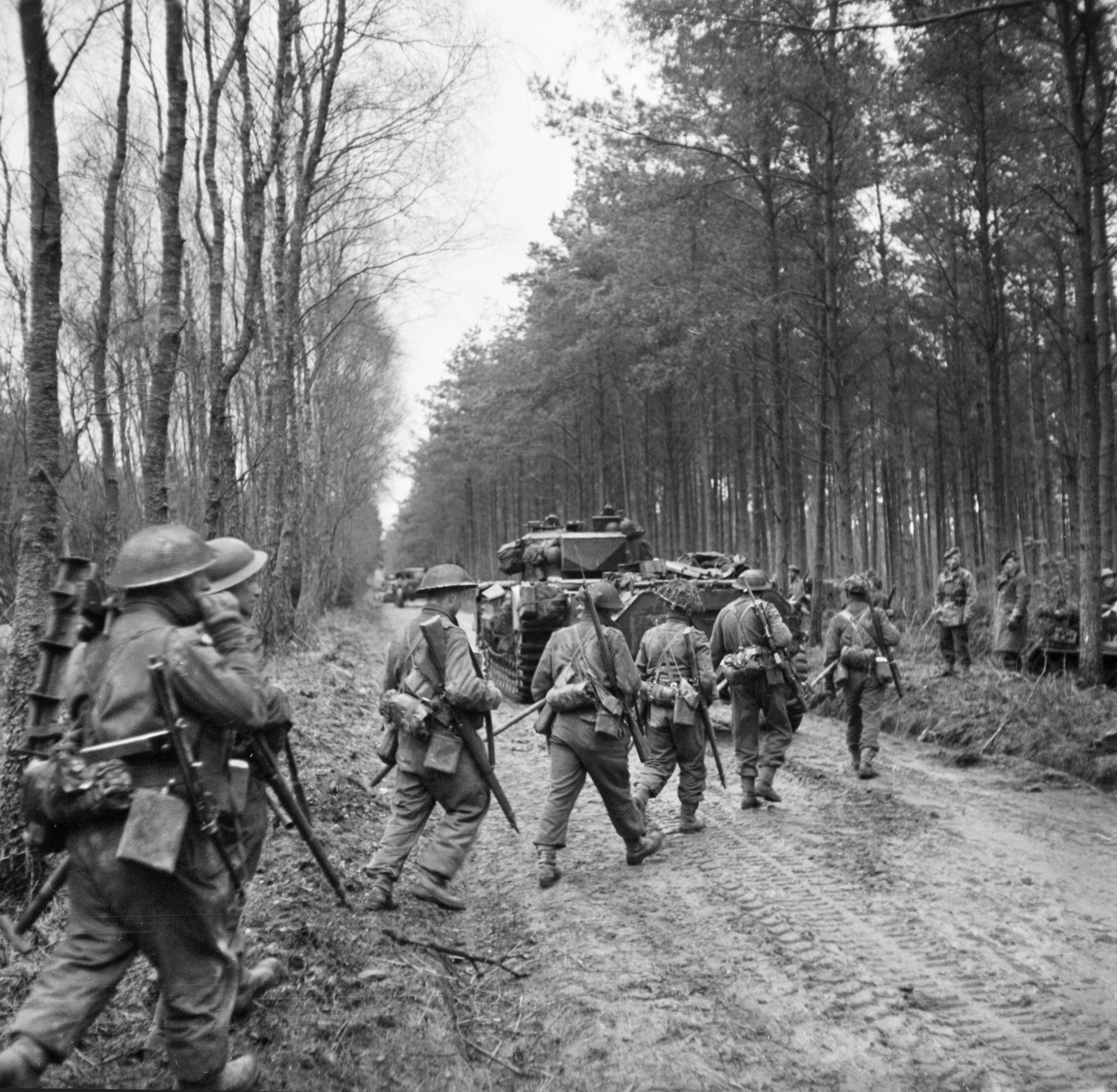 British soldiers of the 2nd Seaforth Highlanders march along a muddy road in the Reichswald accompanied by a supporting medium tank. 