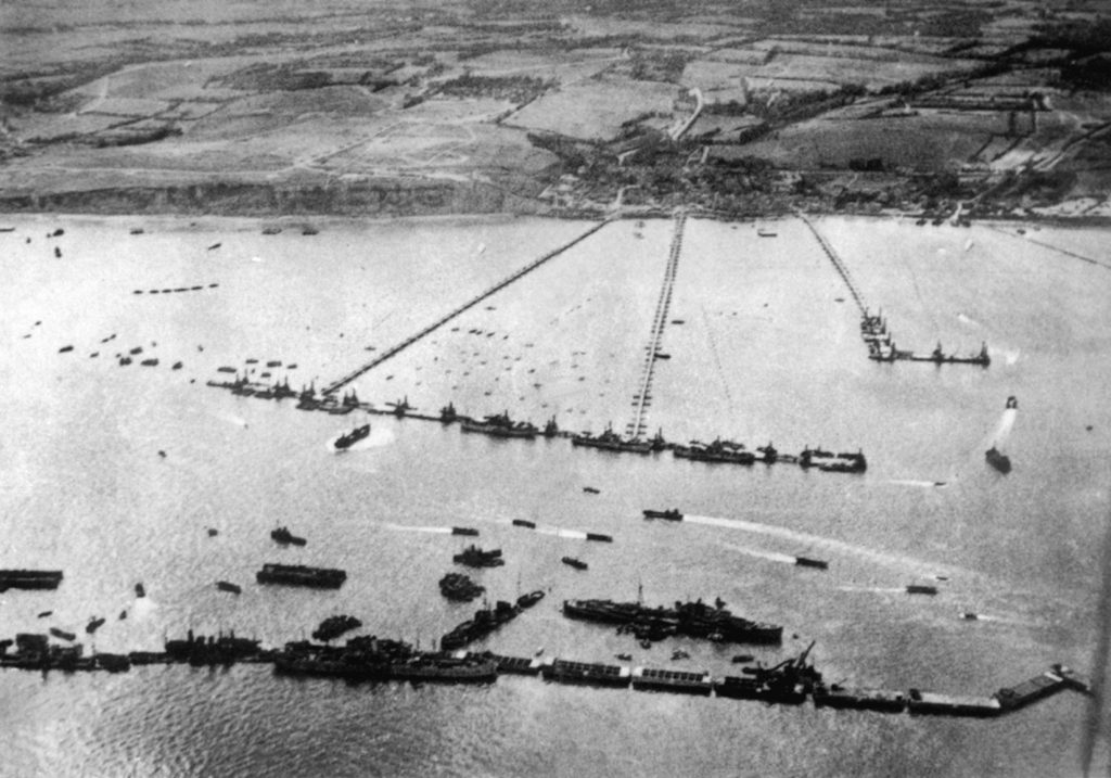 This aerial view depicts a Mulberry harbor completely deployed off the coast of Normandy and in full operation. The breakwater consisting of caissons and block ships is shown at bottom, while the row of pier heads forms a wharf at center, and the floating roadways run directly toward the beach. When fully assembled, a Mulberry harbor was roughly the same size as that of Dover, England.