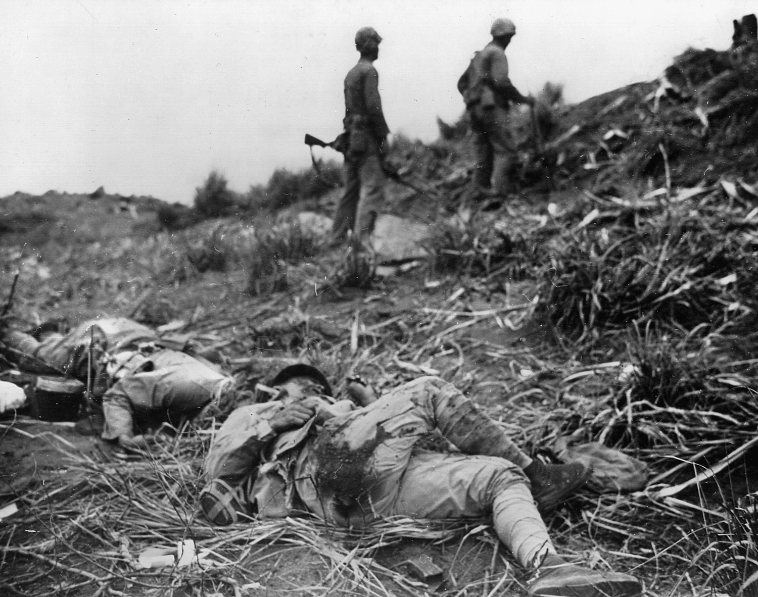 Japanese dead litter the ground near two Marines warily making their way up a hill toward their next objective. 