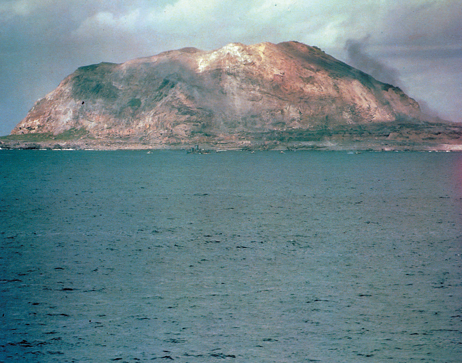 The 554-foot-tall Mount Suribachi looms in the background as the wakes of landing craft carrying invading U.S. troops stretch like fingers in the waters off the coast of Iwo Jima.